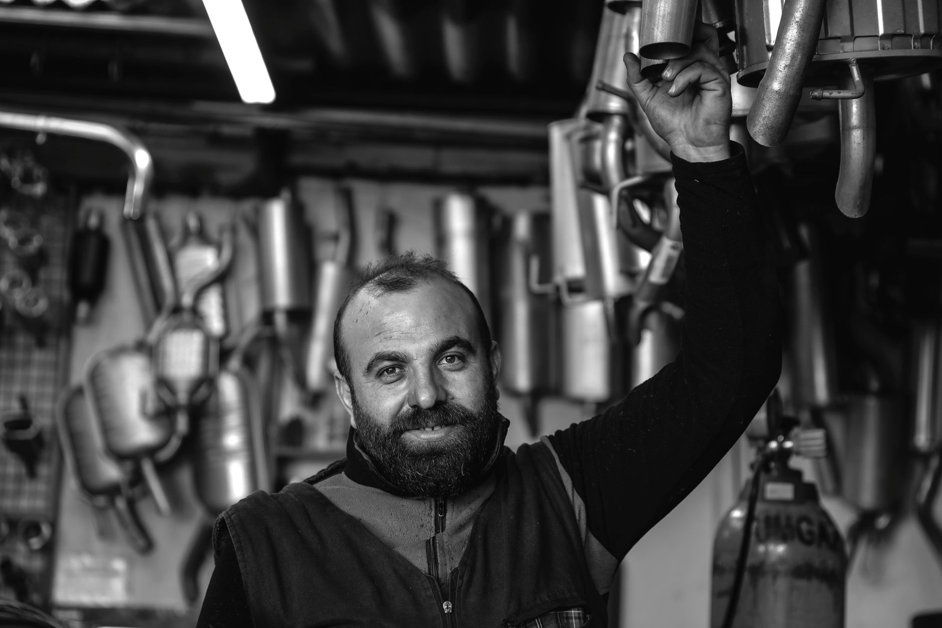 Mecánico sonriente sosteniendo una pieza de escape, rodeado de repuestos en su taller
